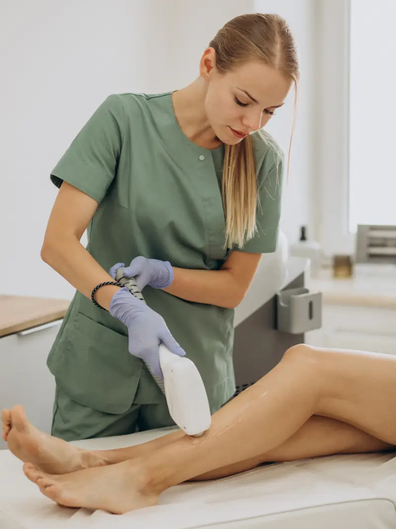 home page 3rd location A technician in green scrubs uses a laser device on a client's leg during a hair removal treatment in a bright clinic setting.