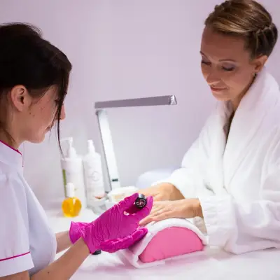 A nail technician in pink gloves uses a tool on a client's hand during a manicure session, set in a modern salon environment.